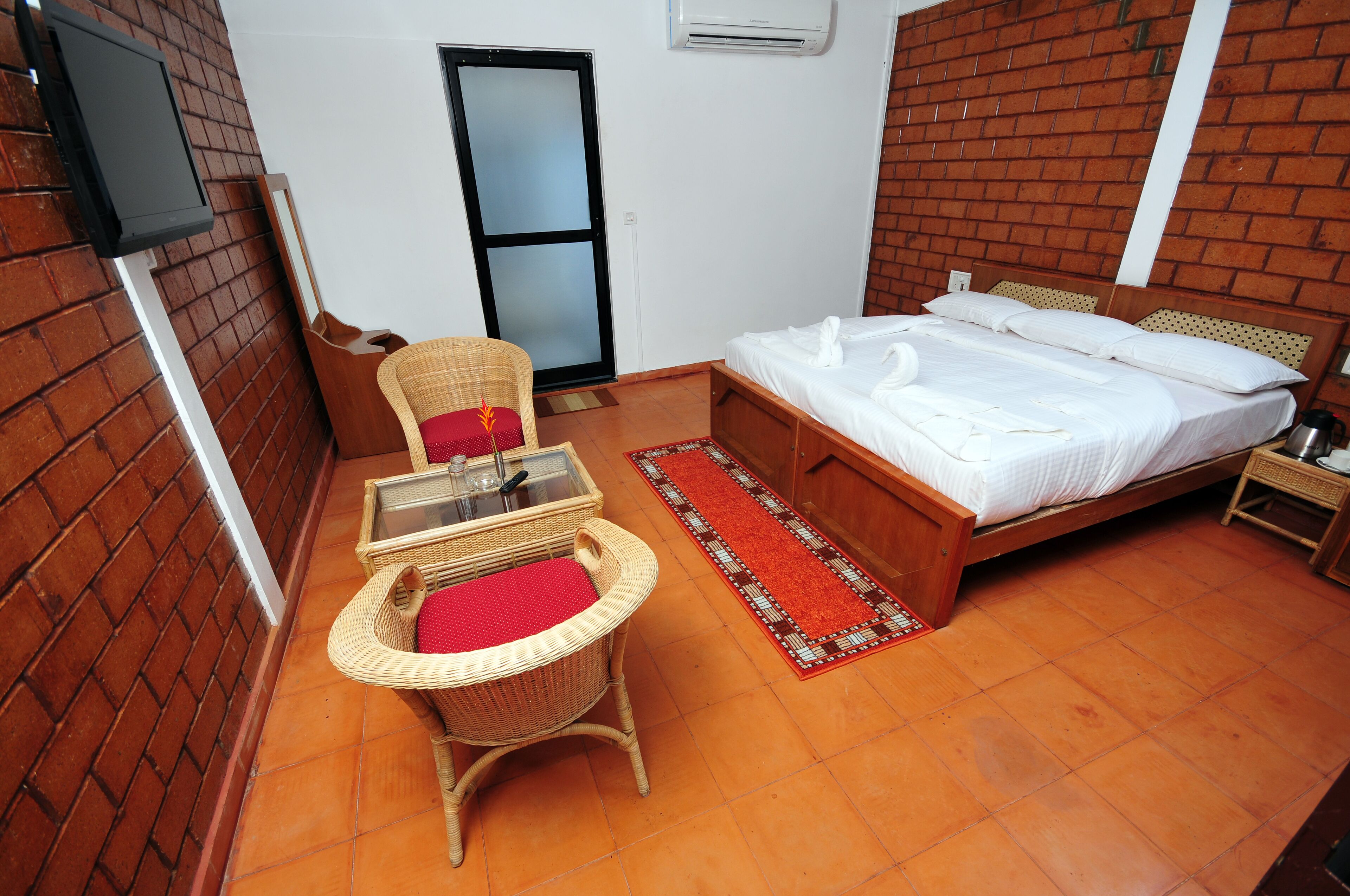 Una habitación con cama, una silla de mimbre con cojín rojo, una mesita con superficie de vidrio, un tapete de diseño rojo y blanco, y una pared de ladrillo.