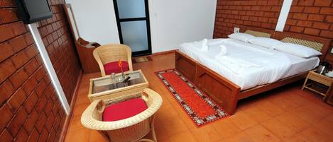 A room with a bed, a wicker chair with a red cushion, a small table with a glass top, a red and white patterned rug, and a brick wall.