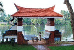 A traditional pavilion with a tiled roof, a small table, and benches, situated by a calm lake with lush greenery in the background. - The Lake Resort by Maat Hotels (Kuttanad Taluk)