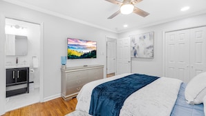 A bedroom with a bed, a ceiling fan, a dresser, a vanity, and a large window.