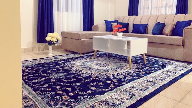 A living room with a beige sofa, blue and white curtains, a white coffee table, and a patterned rug. - Lux Suites Polytech Court Apartments (Nairobi)