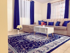 A living room with a beige sofa, blue and white curtains, a white coffee table, and a patterned rug.