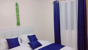 A bedroom with a bed, white headboard, blue bedding, and a curtain. - Lux Suites Polytech Court Apartments (Nairobi)