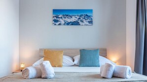 Een netjes opgemaakt bed met twee opgerolde handdoeken, een houten hoofdbord en een schilderij van een berglandschap erboven.