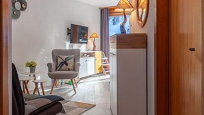 Een moderne woonkamer met een bank, fauteuil en een kleine tafel met een plant. Er is een televisie aan de muur bevestigd, een lamp en een houten kast.
