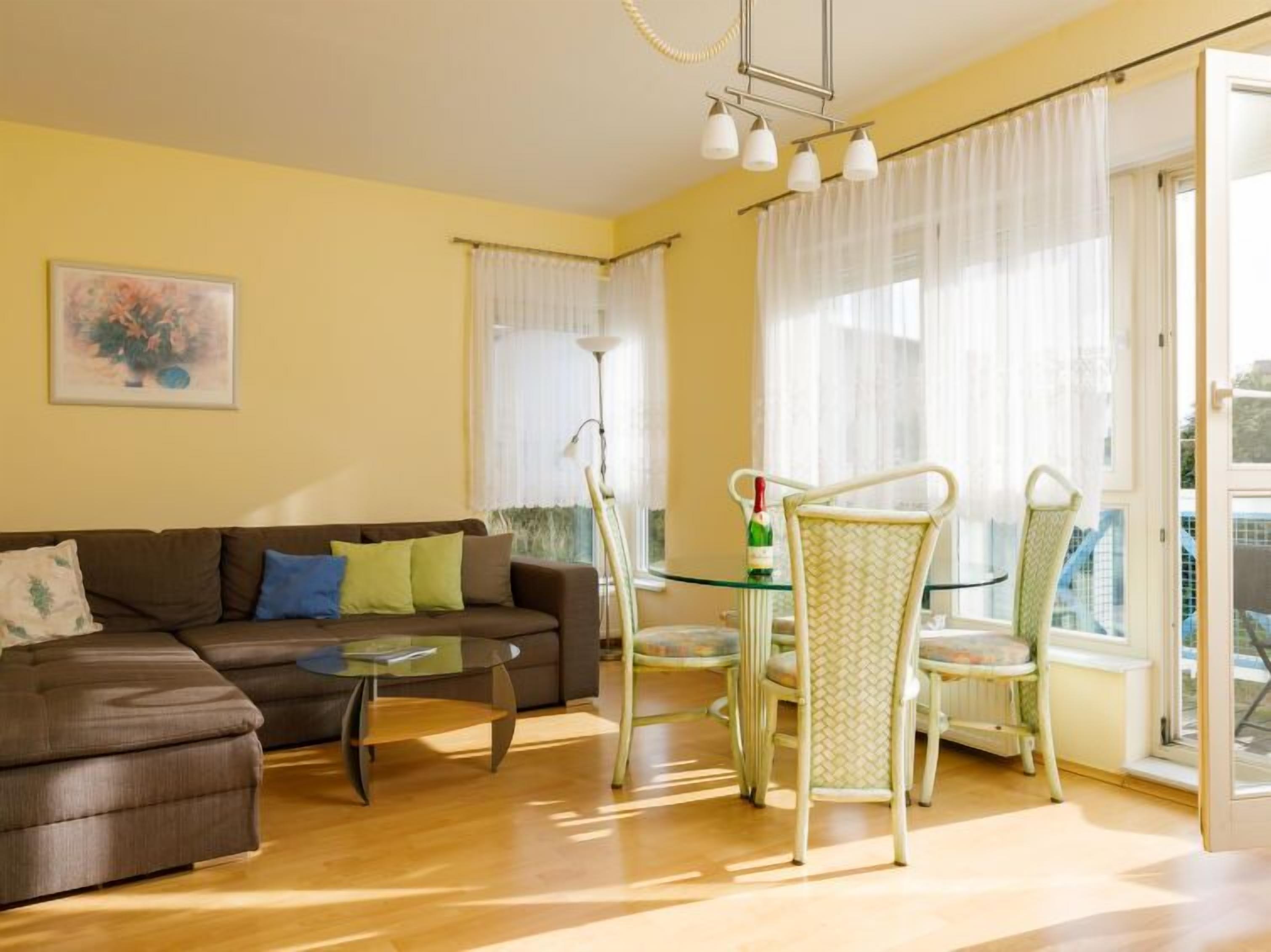 A living room with a brown sofa, a glass coffee table, and a dining area with a glass table and chairs. There is a painting on the wall and a bottle on the table.