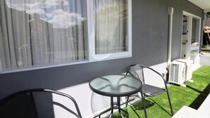 A balcony with a glass table and chairs, a grey wall, and a window with curtains. - Barnes Street BNB - Enjoy your stay in lovely private unit.
 (Timaru)