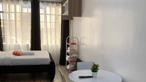 A modern hotel room with a bed, a round dining table with chairs, a small potted plant, and a metal shelf unit.