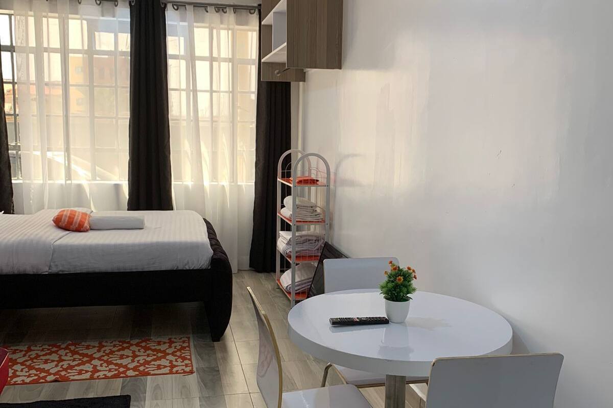 Una habitación de hotel moderna con una cama, una mesa redonda de comedor con sillas, una pequeña planta en maceta y un estante metálico.