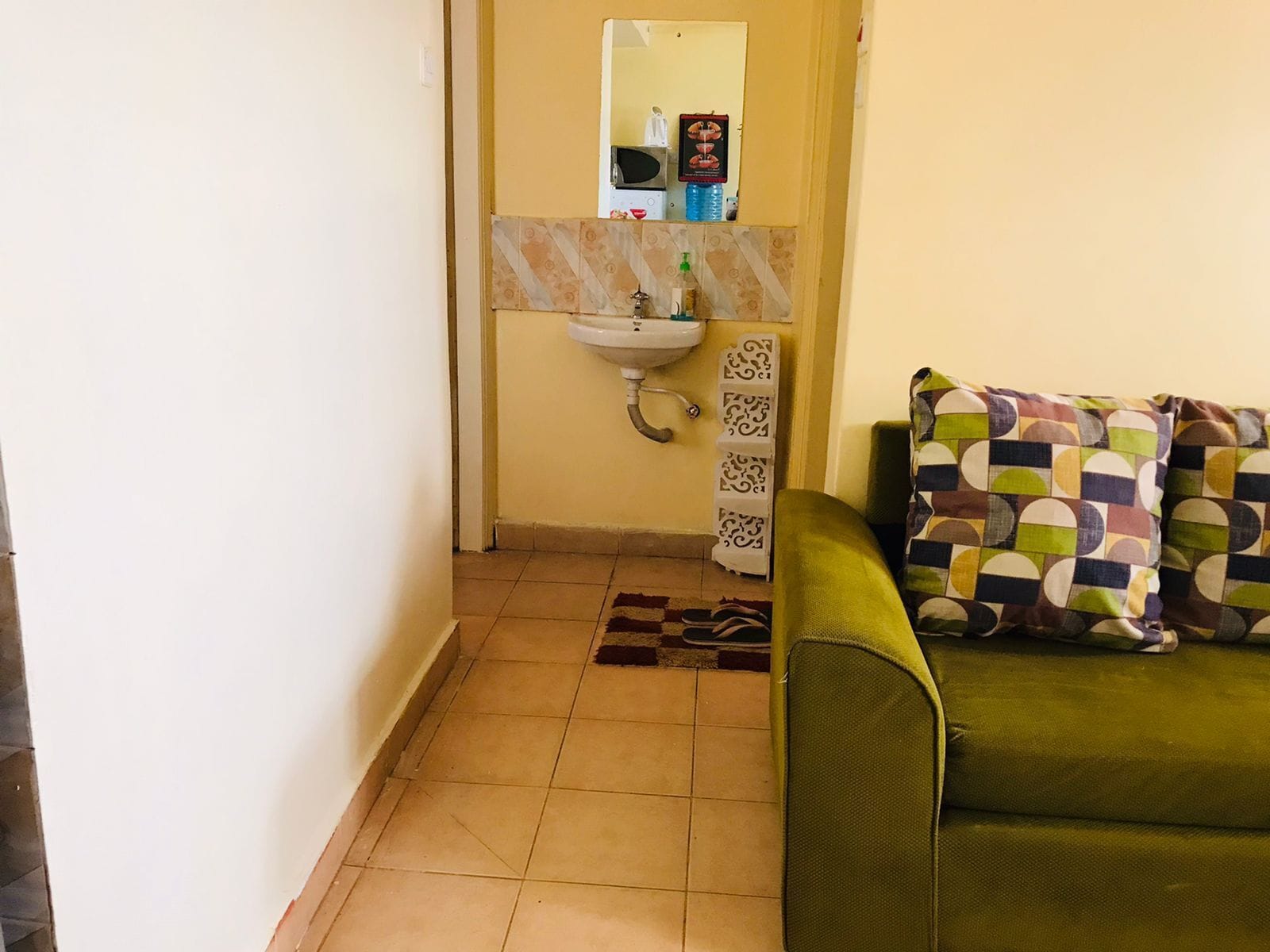 A small bathroom with a sink, mirror, and patterned tiles. There is a green sofa with a colorful cushion.