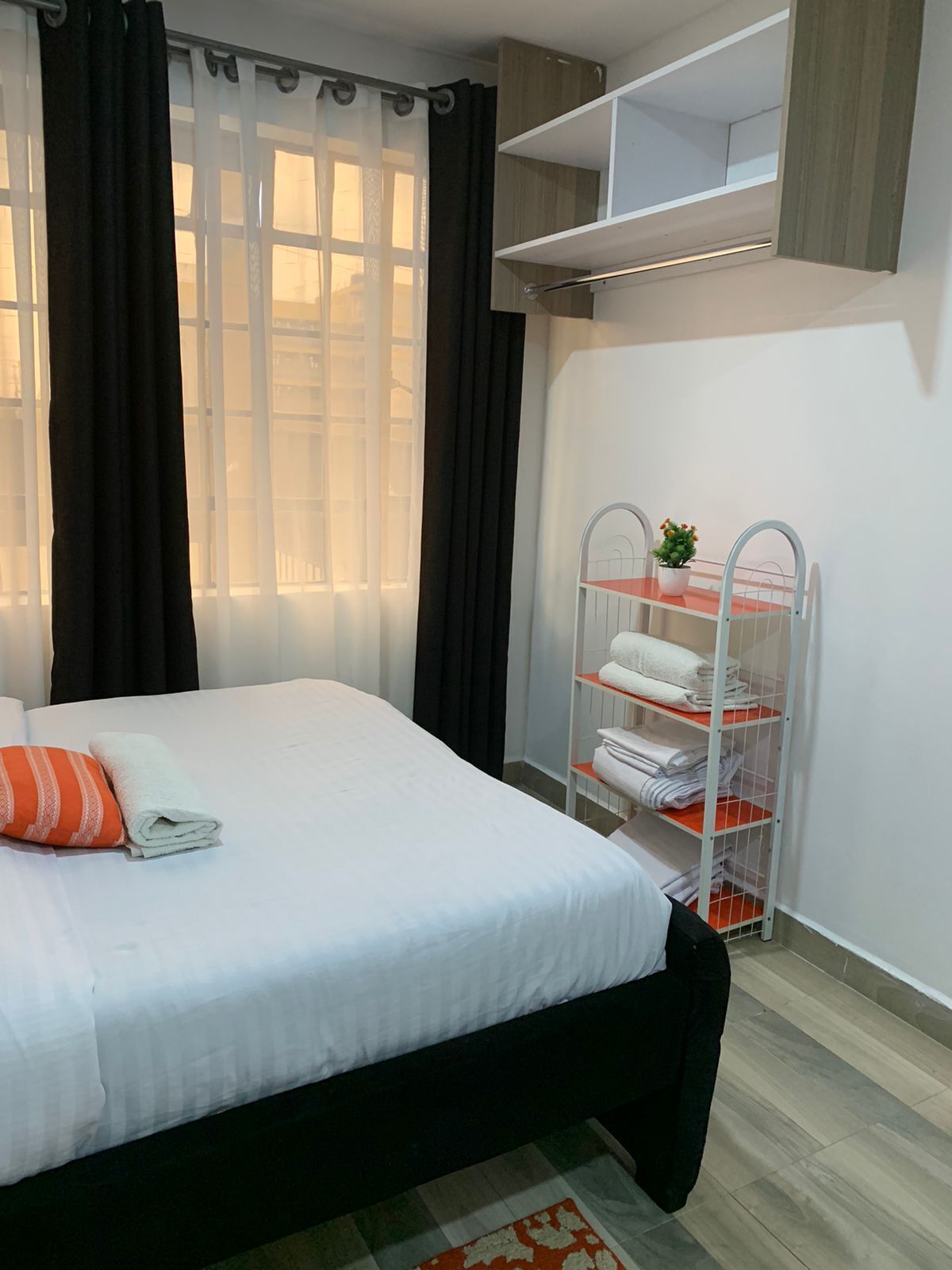 A neatly made bed with a white comforter and an orange pillow, a window with sheer curtains, a metal shelf unit with folded towels, and a small potted plant.