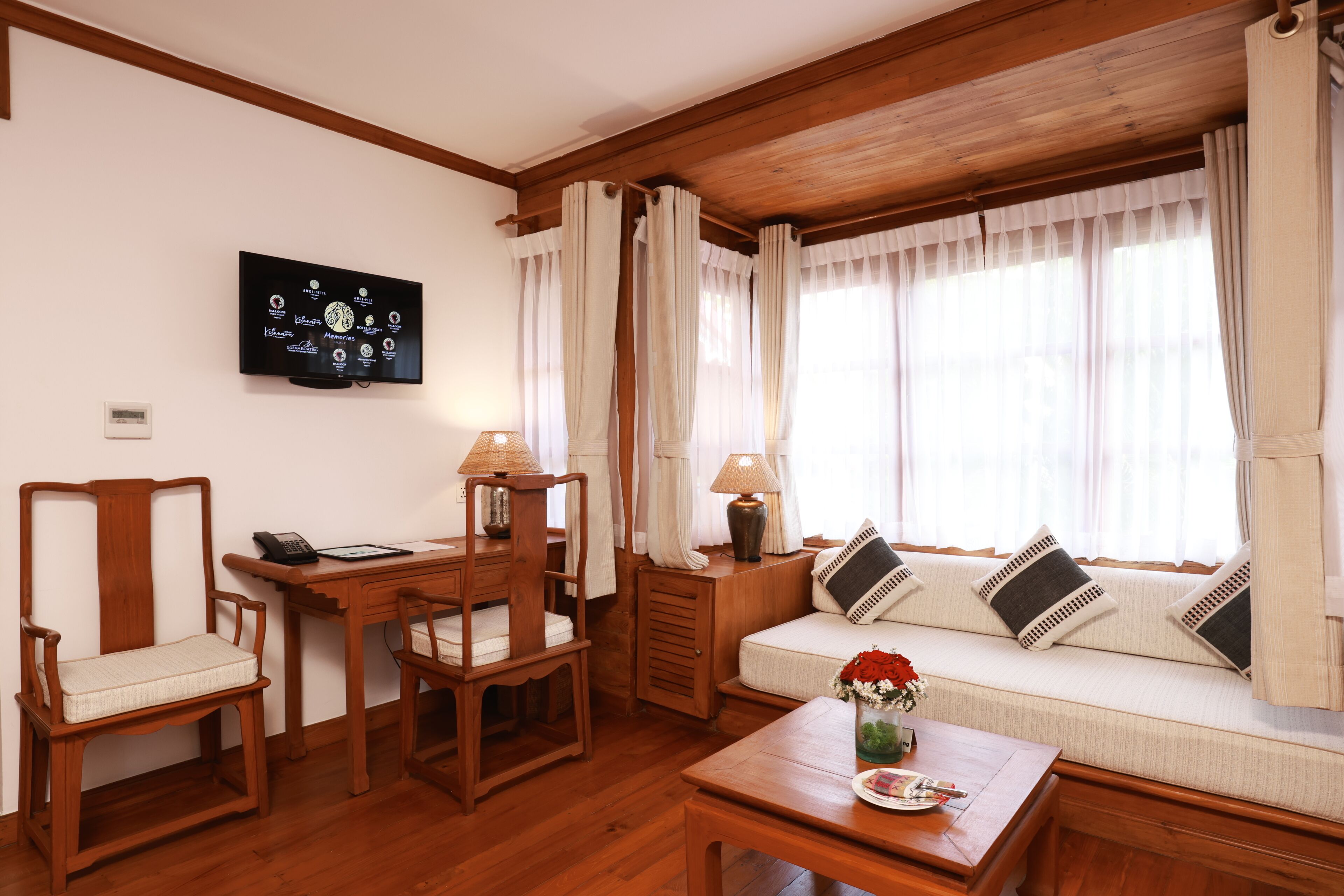 A cozy living room with a wooden floor, a sofa with cushions, a coffee table with a vase, a desk with a lamp, and a television mounted on the wall.