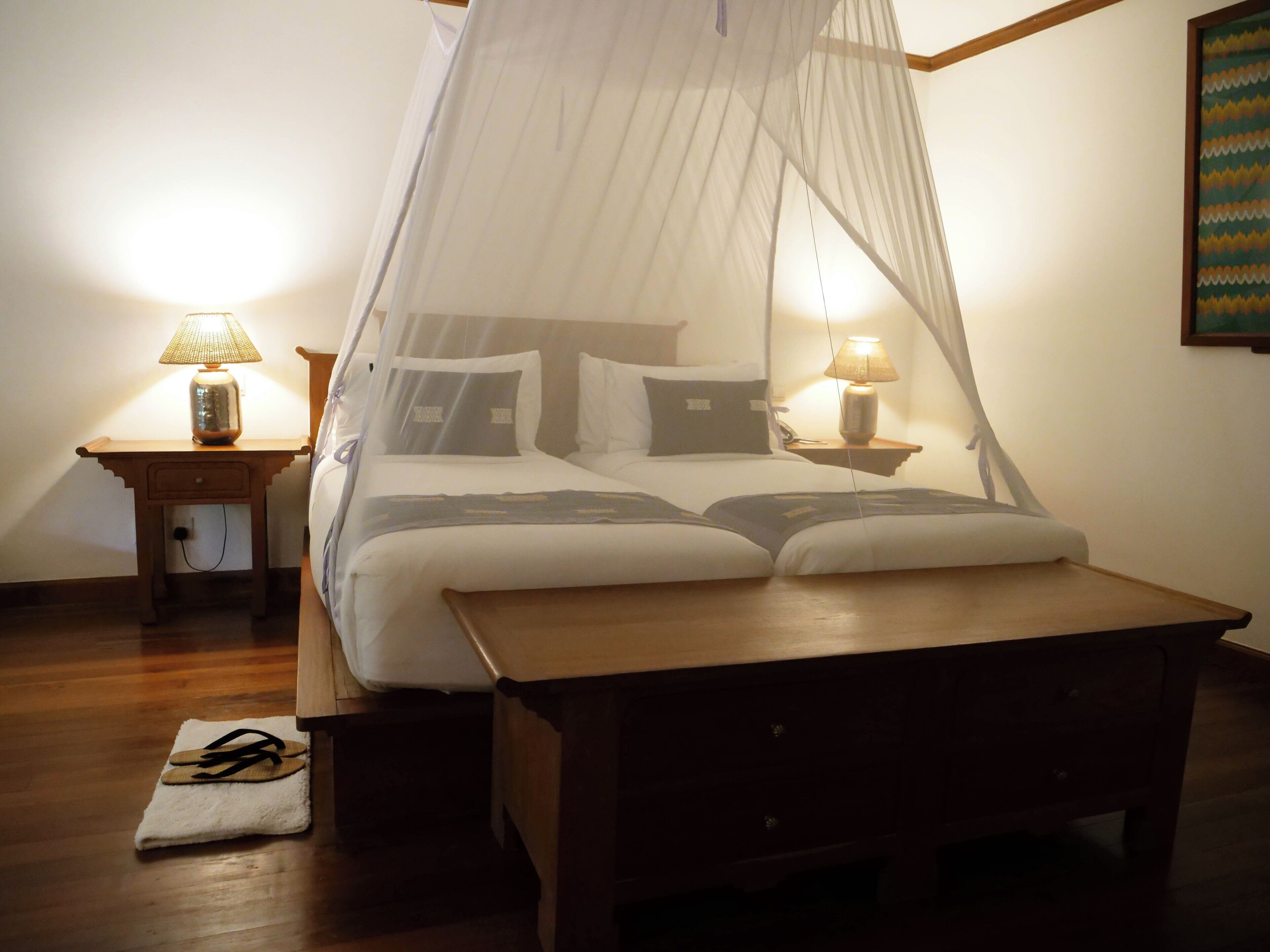 A bedroom with a canopy bed, two bedside tables with lamps, a wooden nightstand, and a pair of flip-flops on a towel.