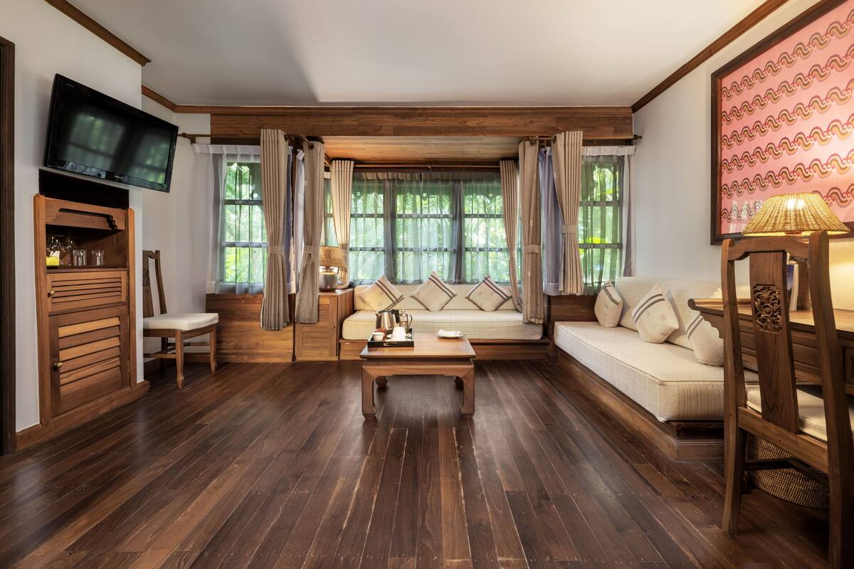 A cozy living room with a wooden floor, a sofa, a coffee table, a TV mounted on the wall, and a window with curtains.