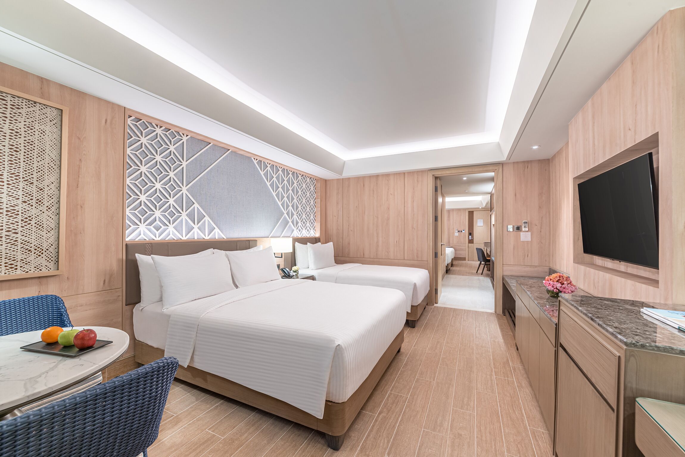 Quarto de hotel com uma cama grande, duas mesas de cabeceira, uma TV e um espaço de refeições com cadeiras de vime.