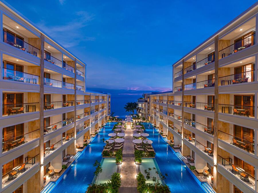 A multi-story hotel with a central swimming pool, balconies with seating, and a view of the ocean.