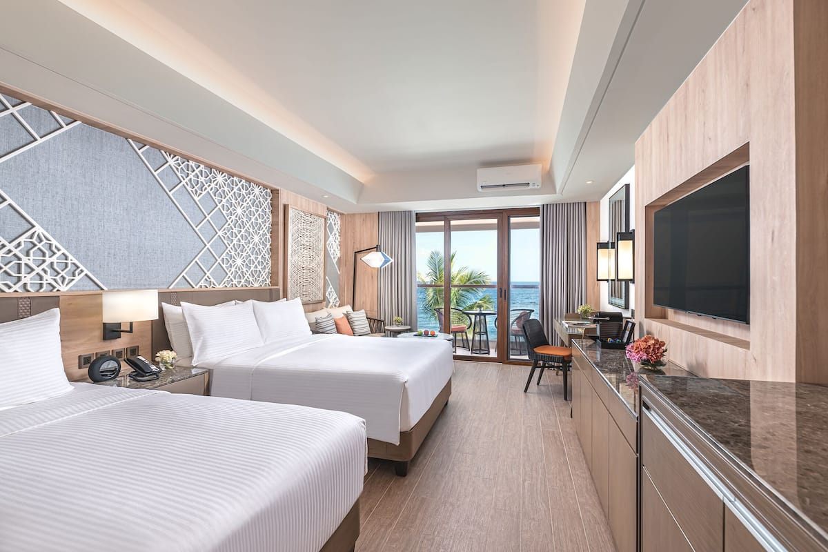 A hotel room with two beds, a desk with a chair, a TV, and a balcony with a view of the ocean.
