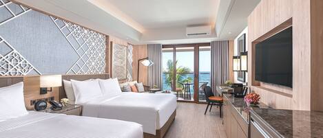 A hotel room with two beds, a desk with a chair, a TV, and a balcony with a view of the ocean.