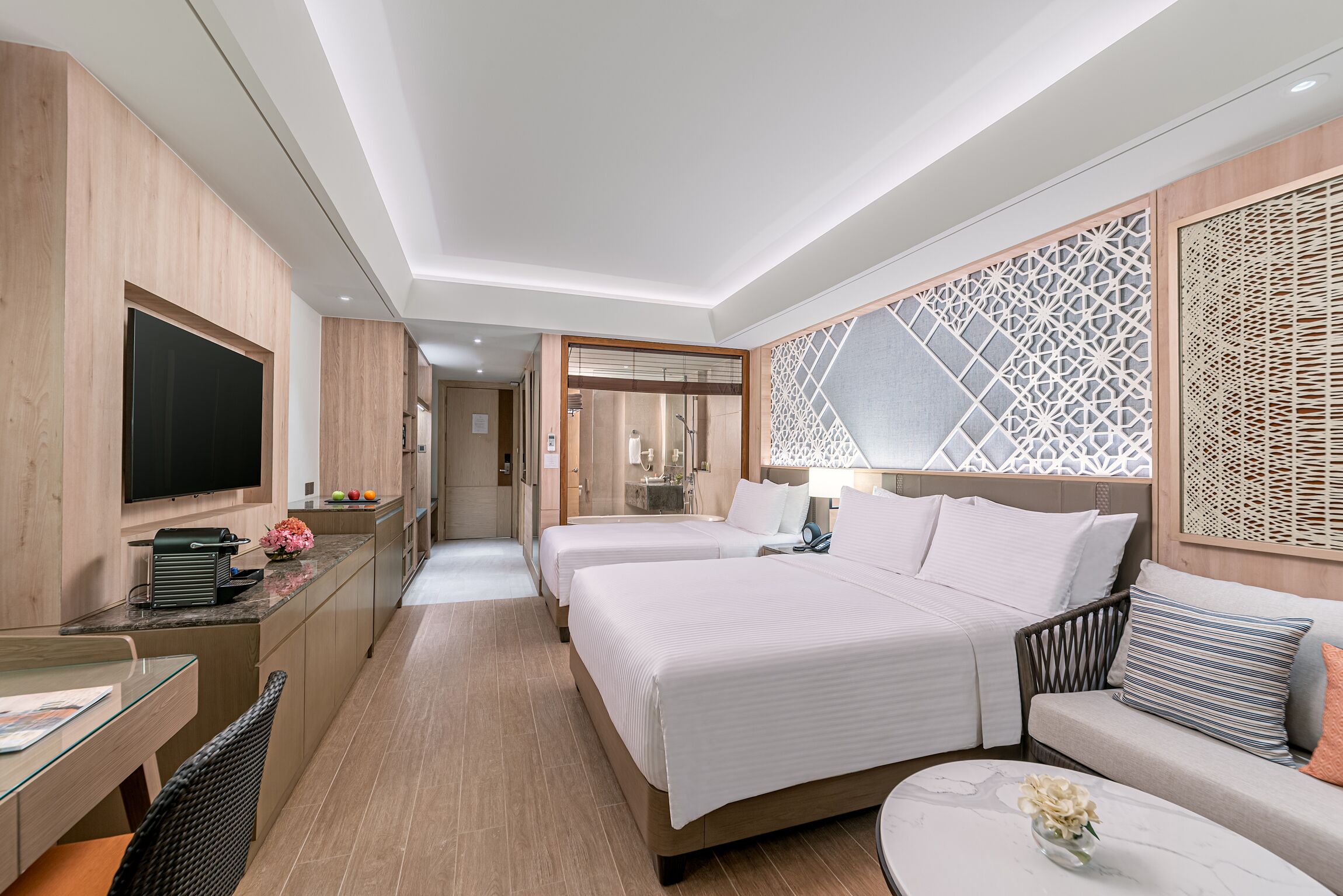 A modern hotel room with two beds, a desk with a flat-screen TV, a sofa, and a decorative wall panel.