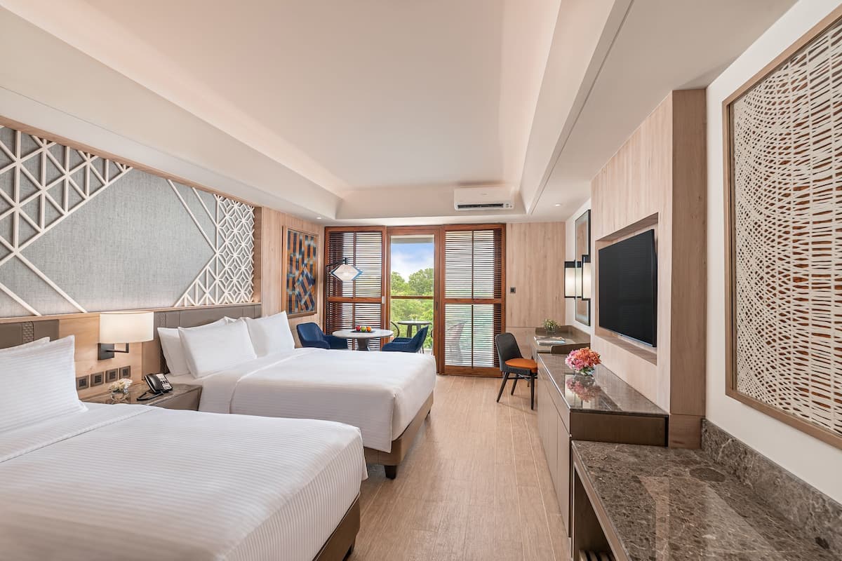 A hotel room with two beds, a desk with a TV, a small table with chairs, and a balcony with a view.