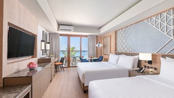 A hotel room with two beds, a TV, a desk, a chair, a small table, and a view of the ocean.