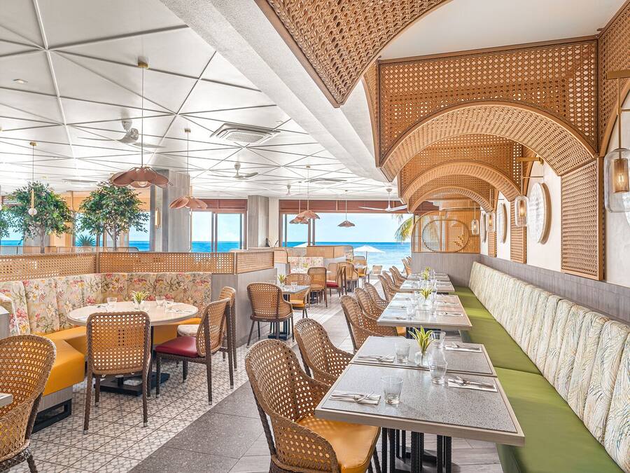 Un restaurante con vista al océano, que cuenta con mobiliario de mimbre, asientos con cojines verdes y un diseño de techo moderno.
