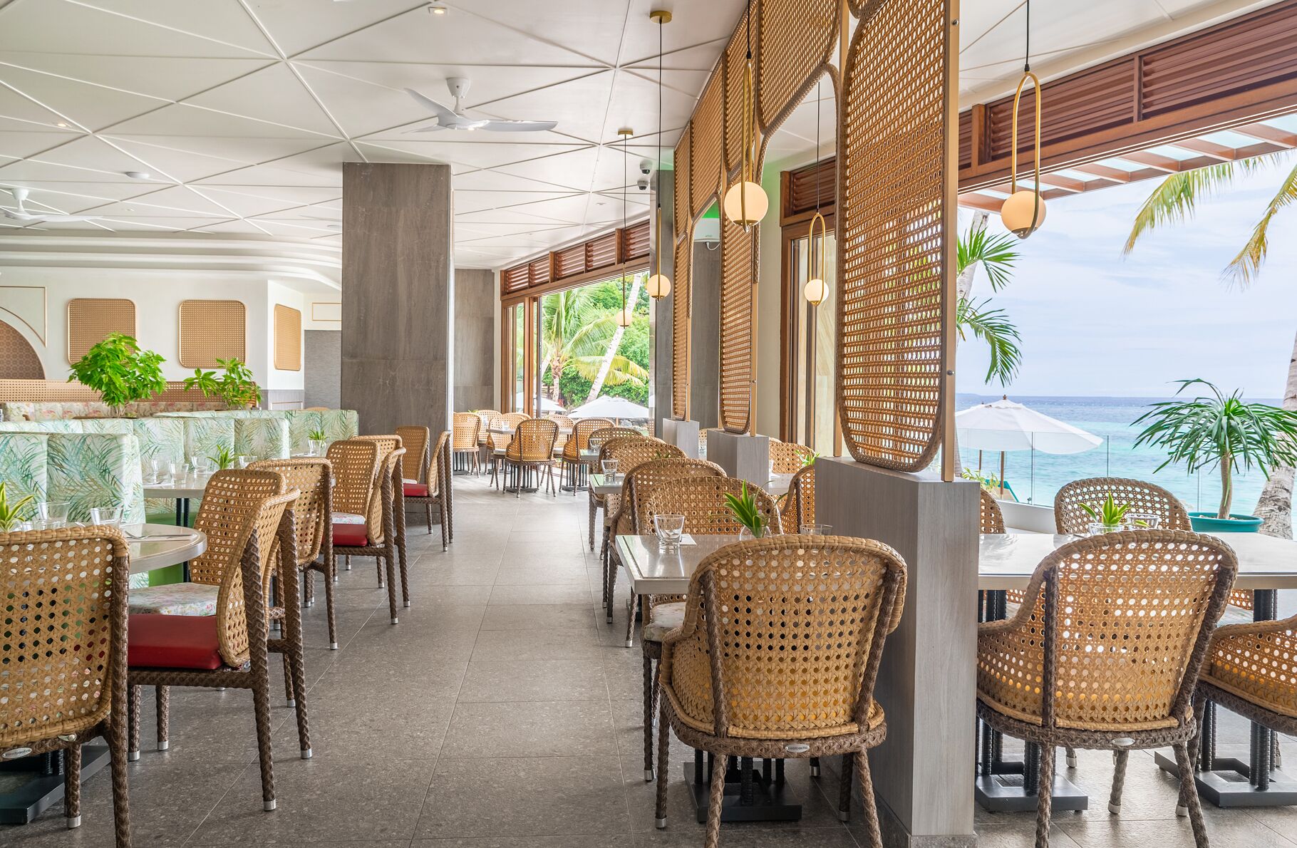 A restaurant with wicker chairs and tables, large windows with ocean views, and a ceiling with geometric patterns.