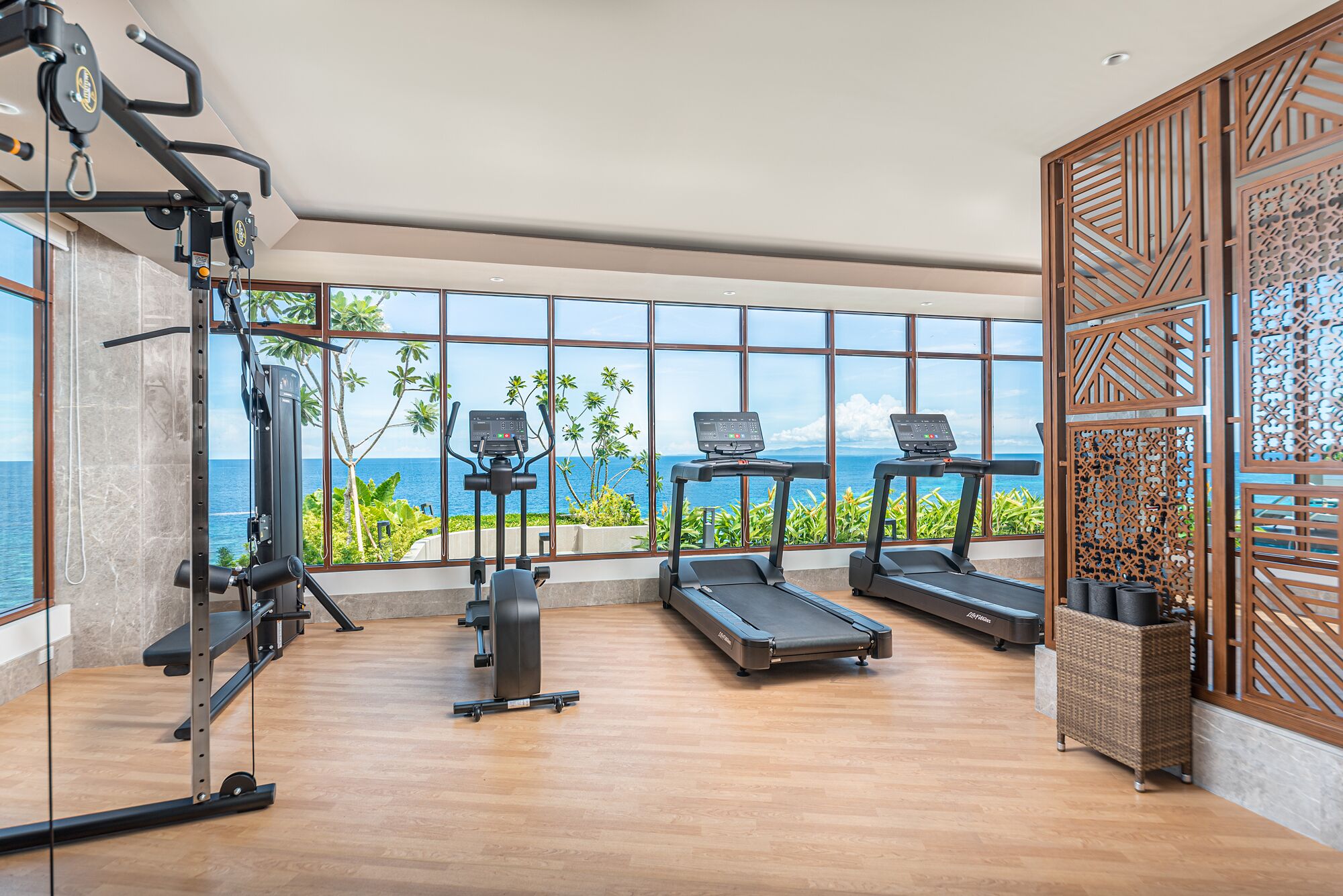 Une salle de sport équipée de matériel d’exercice, de grandes fenêtres offrant une vue sur l’océan et un panneau décoratif en bois.