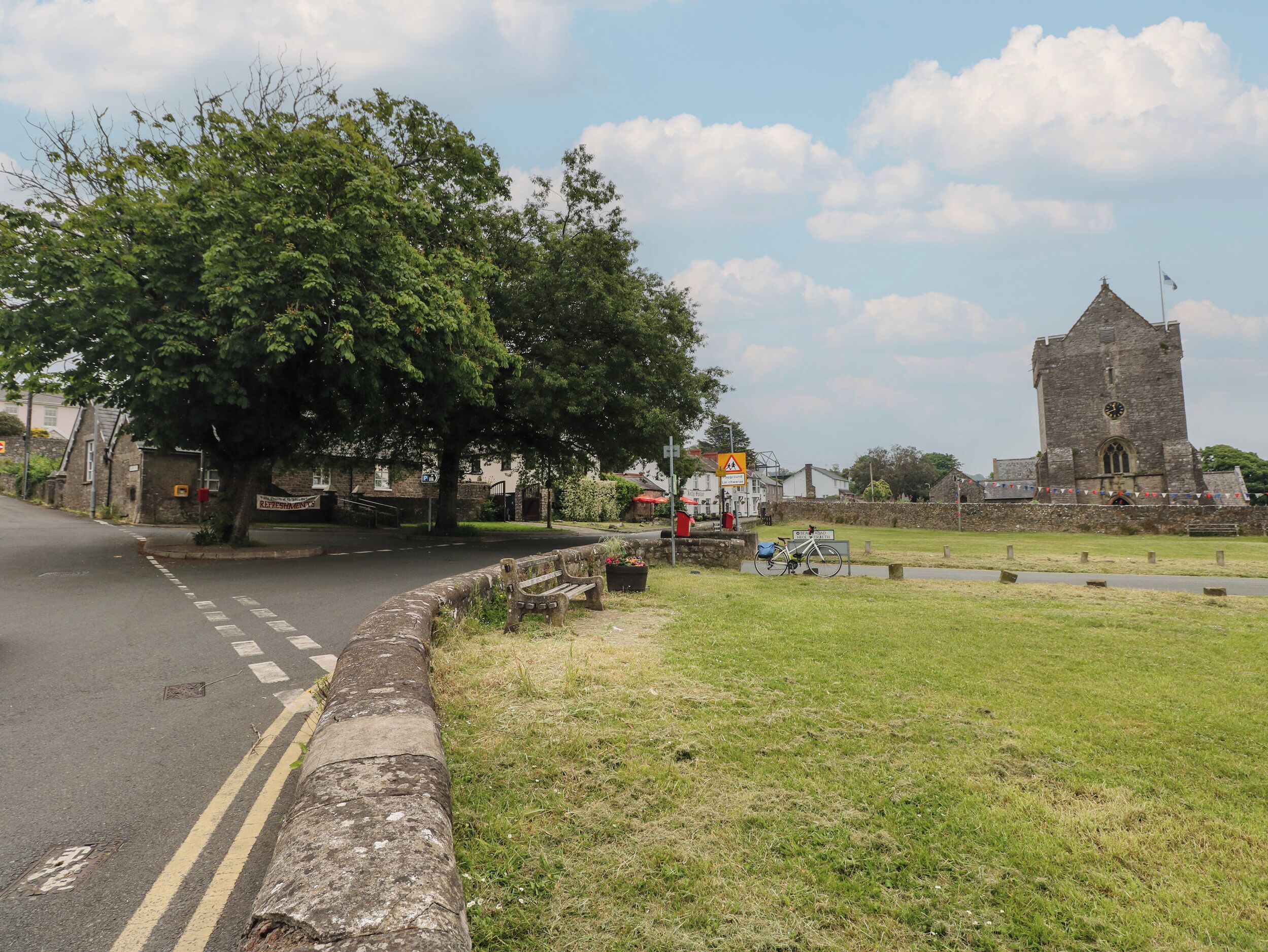 A road curving to the left with a stone wall and a grassy area. A church with a tall tower is visible in the background.