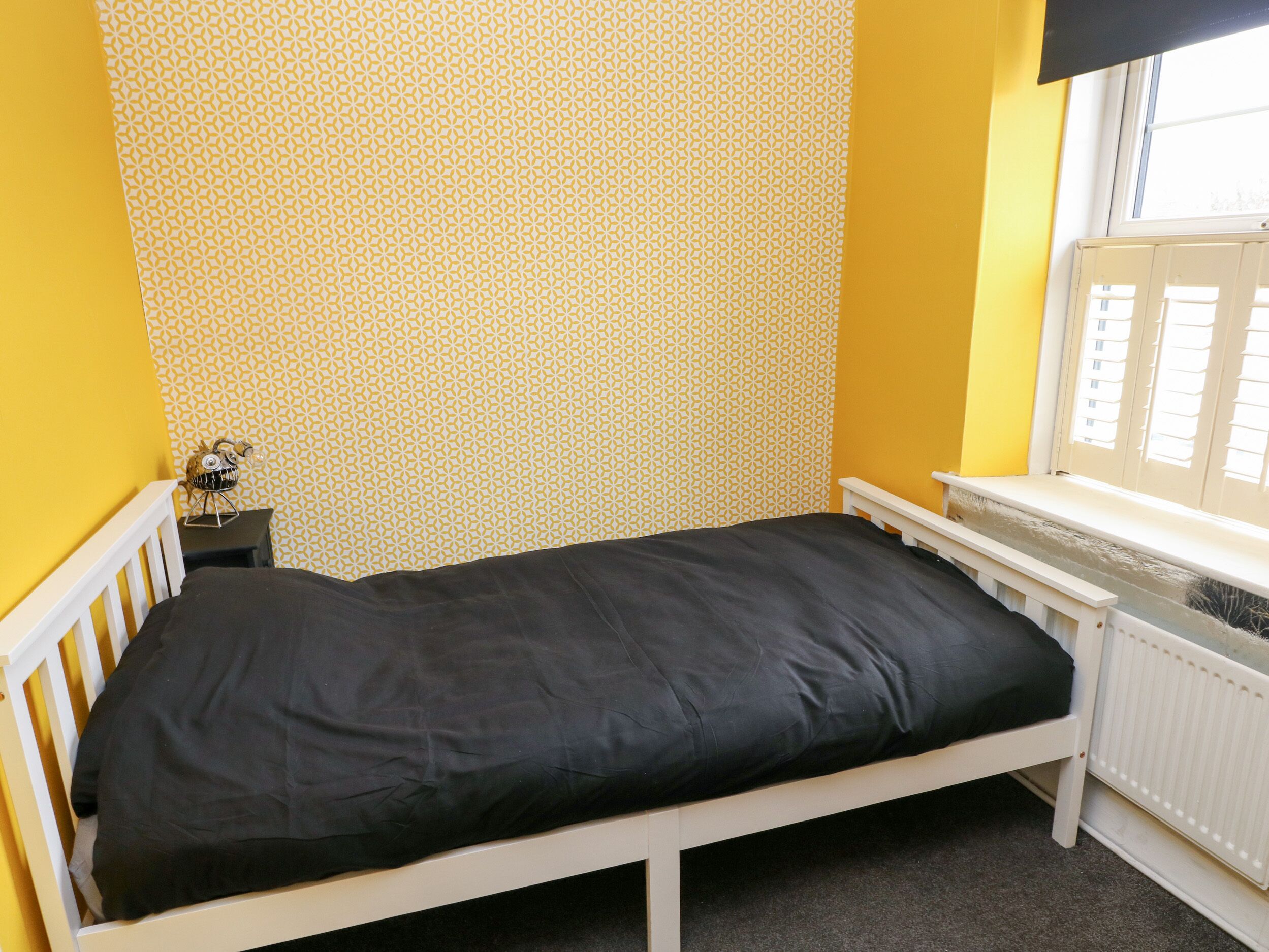 Une petite chambre avec un lit simple, un papier peint à motifs, une fenêtre avec des stores et un radiateur.