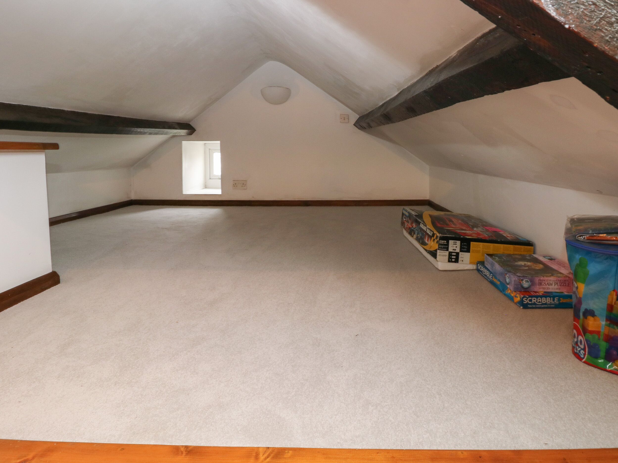 An attic room with a sloped ceiling, a small window, and a wooden beam. There are boxes of games and a container on the floor.