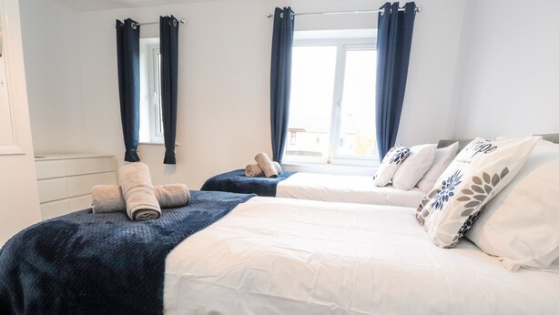 A hotel room with two beds, each with a dark blue bedspread and white linens. There are towels on the beds, and the room has a window with curtains.