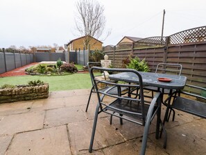 An outdoor patio with a metal table and chairs, a brick planter, and a small garden area with a lawn and a wooden fence. - 3 Thrimby Court (Morecambe)
