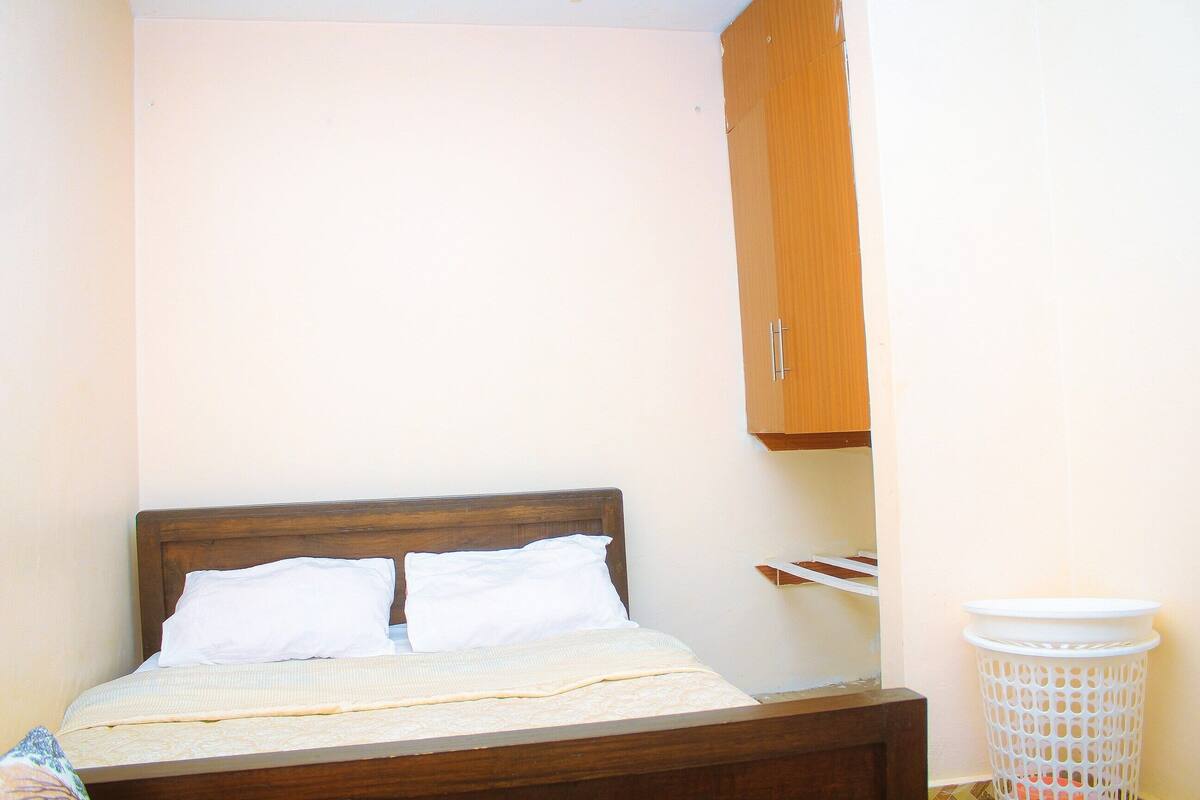 Un lit simple avec une tête de lit en bois, un oreiller blanc et une couverture beige. Au-dessus du lit, on aperçoit une armoire en bois, et à droite, un panier à linge blanc.
