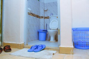 A bathroom with a toilet, a blue trash bin, a blue laundry basket, and a pair of flip-flops on a mat.