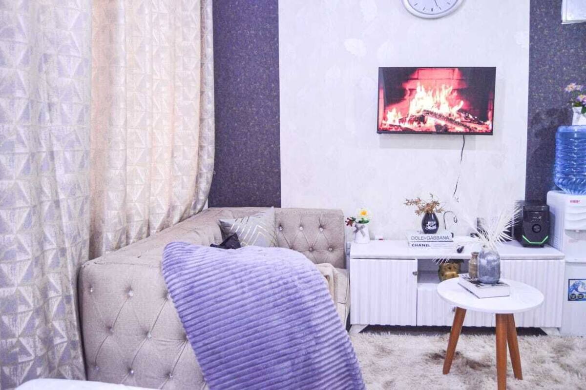 Un salon avec un canapé capitonné, un téléviseur à écran plat, une petite table blanche et un vase décoratif.