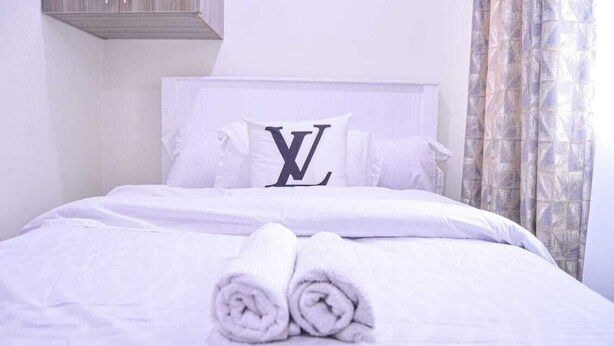 A neatly made bed with white linens, a pillow with a large 'L' logo, and rolled towels on the bed.
