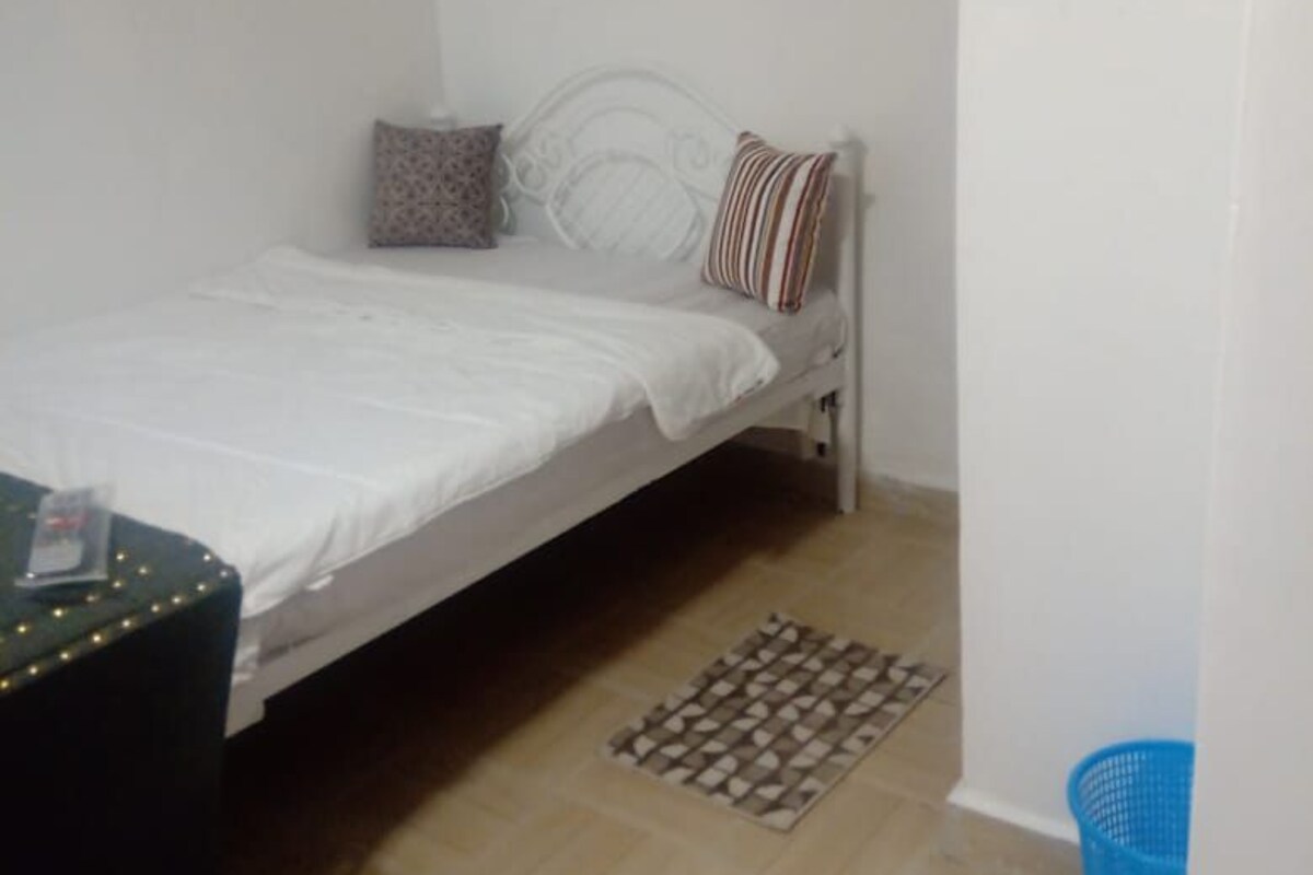 Quarto pequeno e pouco mobiliado com uma cama, uma mesinha e uma cesta azul.