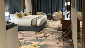 A hotel room with a large bed, a desk, two armchairs, a small table, and a large window with curtains. - Rosa Grand Hotel Monsieh (Riyadh)