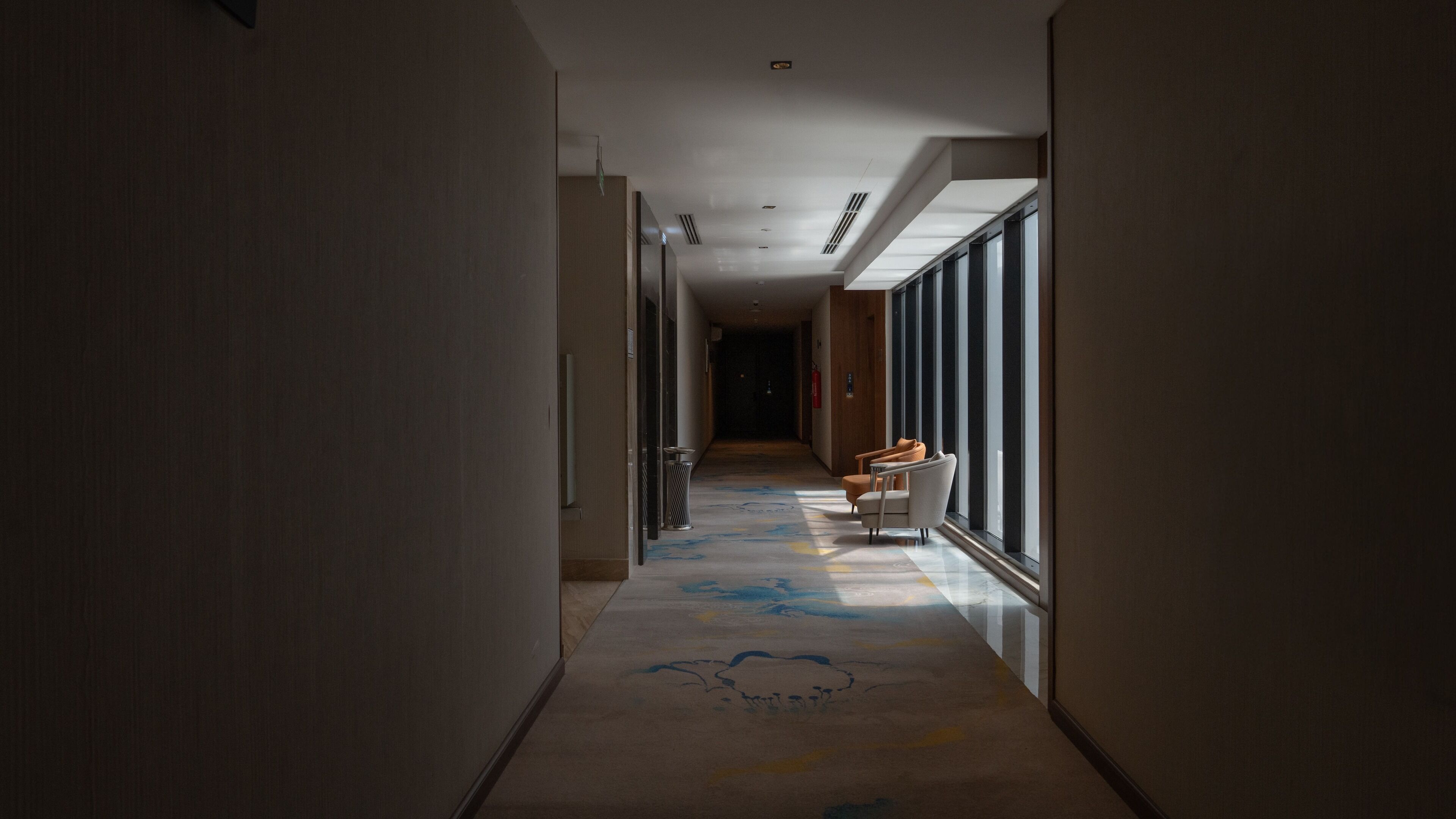 A hotel hallway with a carpet featuring a cloud design, a seating area with chairs, and large windows allowing natural light.