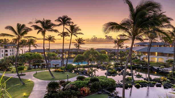 A resort with a pool, palm trees, and a view of the ocean at sunset.