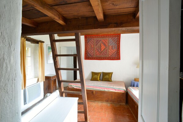 A rustic bedroom with a wooden ladder, a bed with striped bedding, a red patterned tapestry, and a window with curtains.
