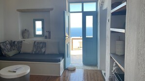 A room with a wooden floor, a blue door leading outside, a white sofa with patterned cushions, and a small round table with a book.