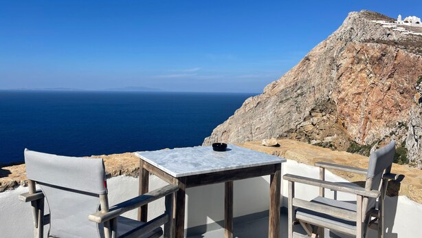 A seaside terrace with a table and chairs, overlooking the sea and a rocky cliff with a white building.