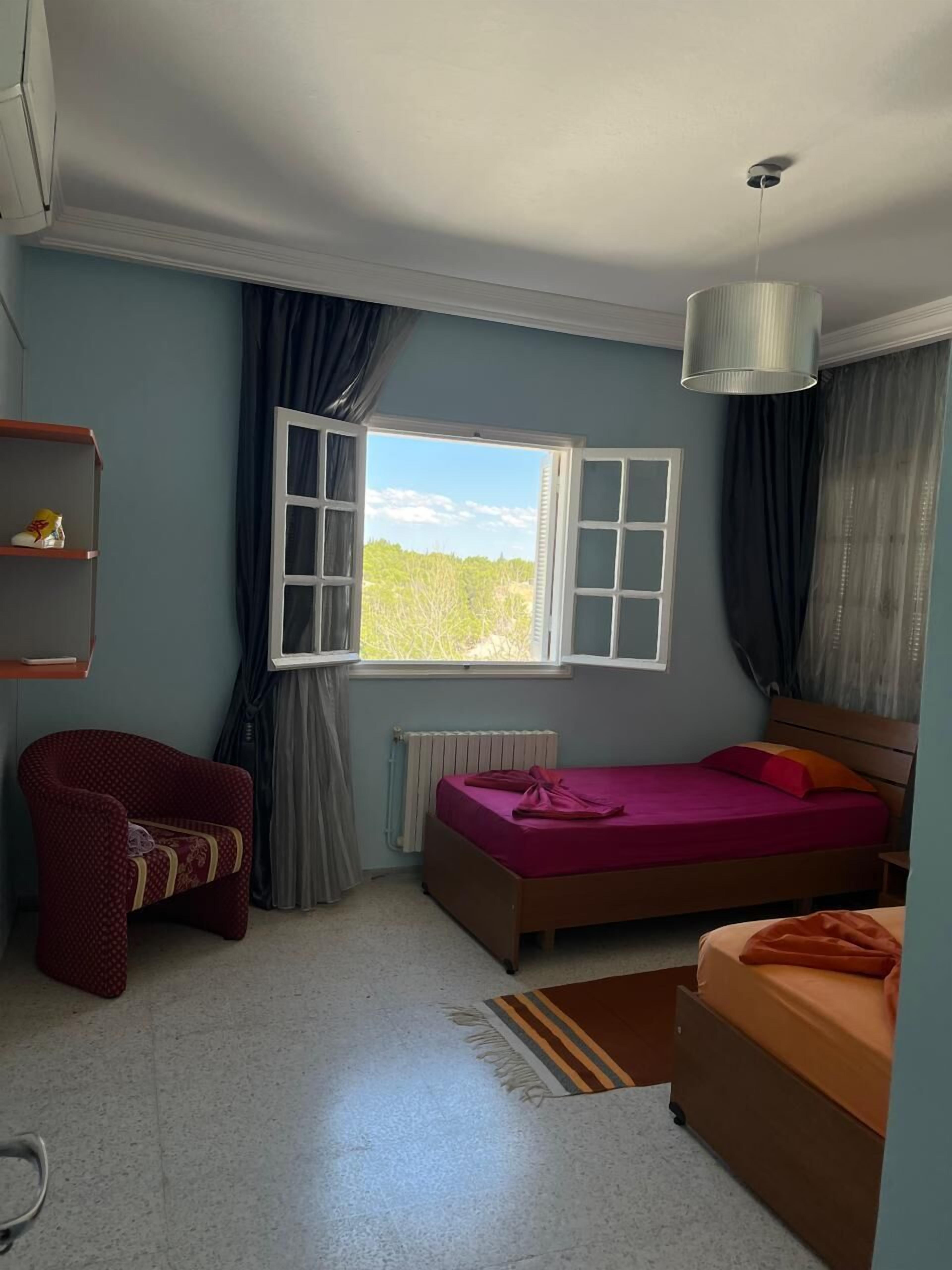 Una habitación con una cama, una silla, un radiador, una ventana con cortinas y una lámpara colgante.