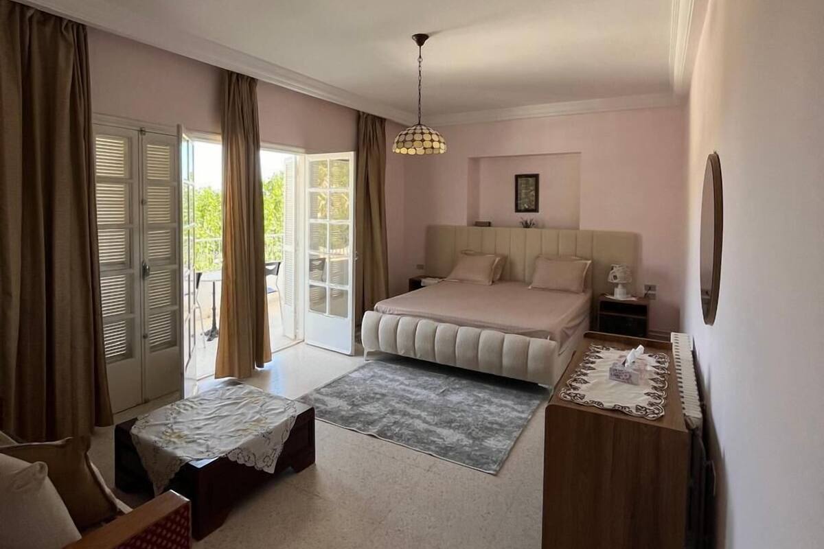 Ein Schlafzimmer mit Bett, Nachttisch, Stuhl, kleinem Tisch, Lampe und Blick ins Freie.