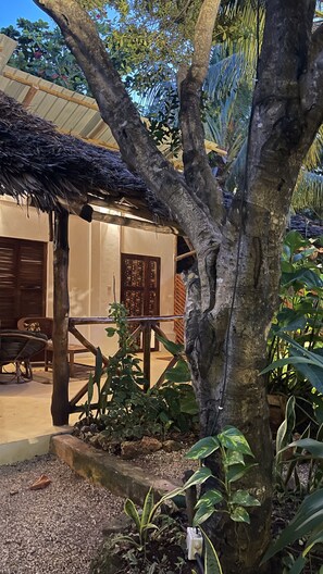 A tree-lined courtyard with a thatched roof structure, a wooden gate, and a building with patterned doors. - Atii Garden Bungalows (Nungwi)