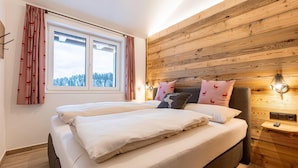 A bedroom with a wooden wall, a large window with curtains, a bed with white bedding, and a nightstand with a lamp. - Holiday Apartment near ski slope (Turracher Höhe)