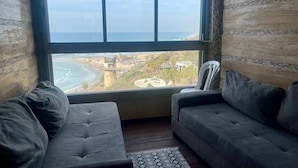 Une pièce dotée d’une grande fenêtre donnant sur une plage et sur le paysage urbain. On y trouve deux canapés de couleur foncée et un fauteuil blanc.