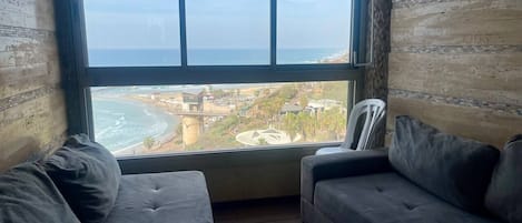 A room with a large window overlooking a beach and cityscape. There are two dark-colored sofas and a white chair.